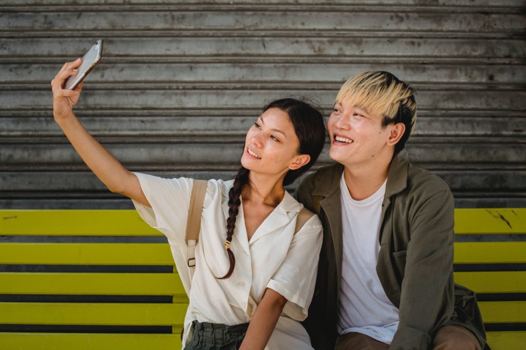 smiling asian couple taking selfie on smartphone on bench