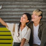 smiling asian couple taking selfie on smartphone on bench