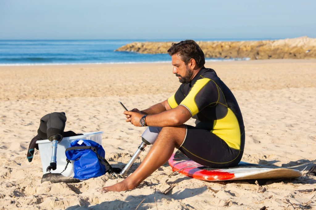 focused ethnic sportsman with leg prosthesis using smartphone on beach